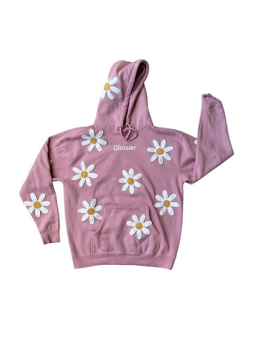 A pink sweatshirt with white daisy appliques and the brand 'Glossier' written across the front.